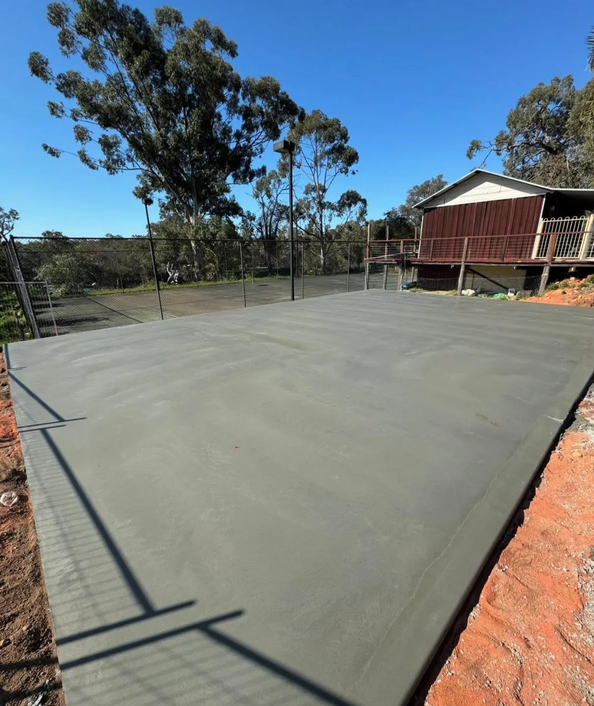 Freshly poured and trowelled grey concrete slab beside a fenced tennis court, with fence shadows falling across the surface