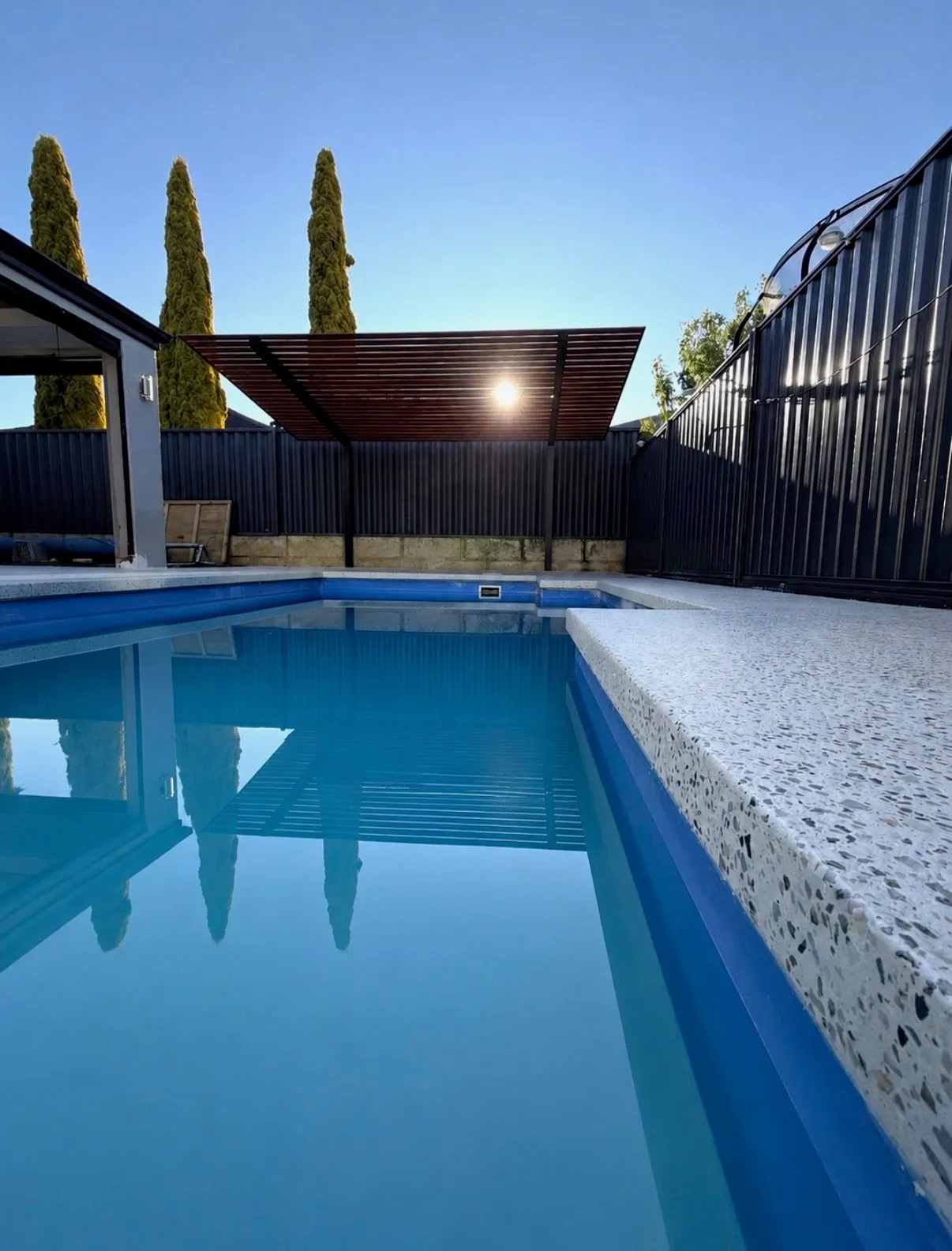 Pale speckled exposed aggregate coping wrapping the edge of a rectangular pool, low sun behind a dark boundary fence