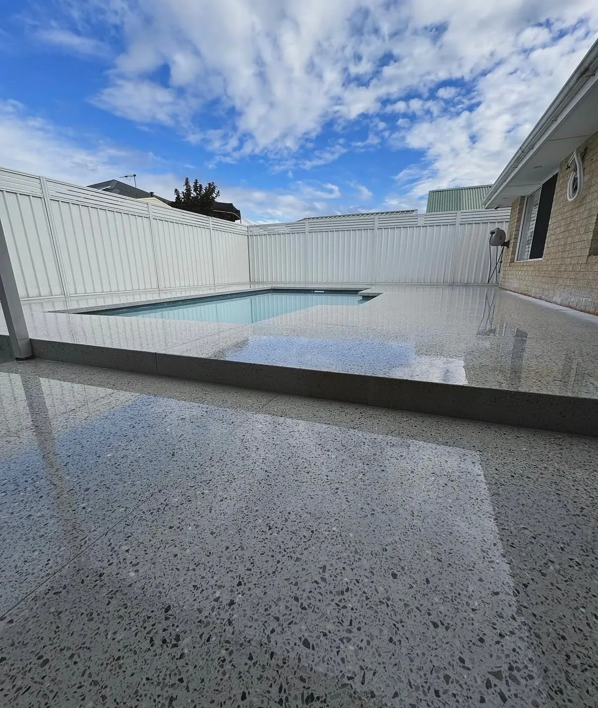 Freshly sealed pale exposed aggregate pool surround, still wet, reflecting the fence and the sky across a wide backyard