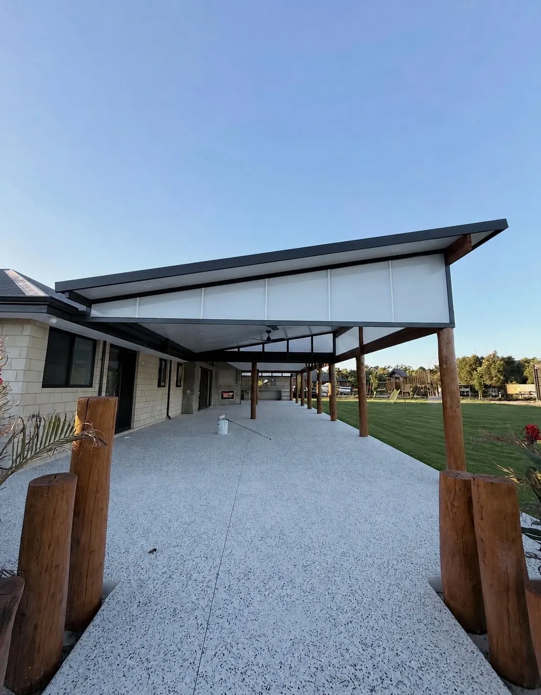 Covered alfresco with a raked roof on timber posts, running out over pale flecked exposed aggregate to a green lawn under a clear sky