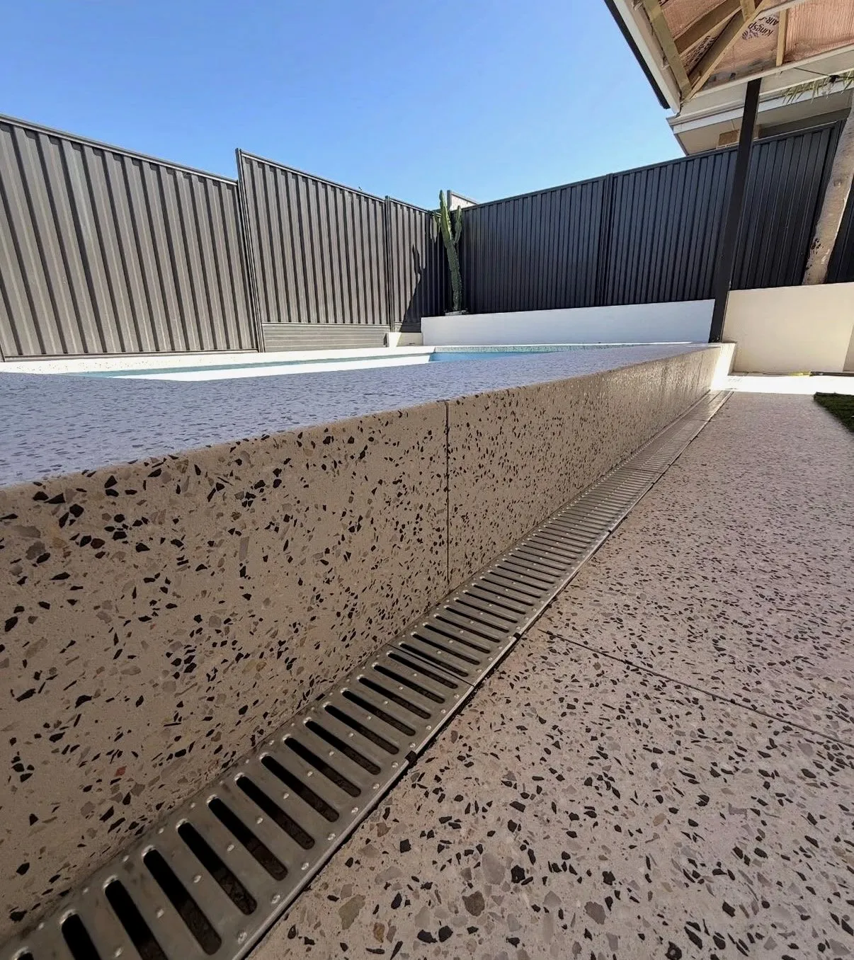 Honed concrete step wall showing a heavy black and white aggregate reveal, with a stainless steel strip drain running along its base