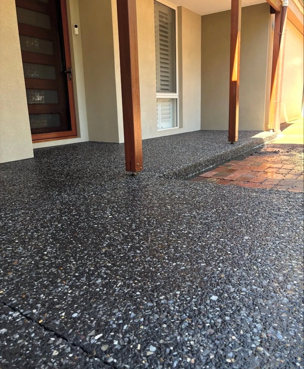 Dark charcoal exposed aggregate verandah and step running past timber posts to a front door, meeting older red brick paving
