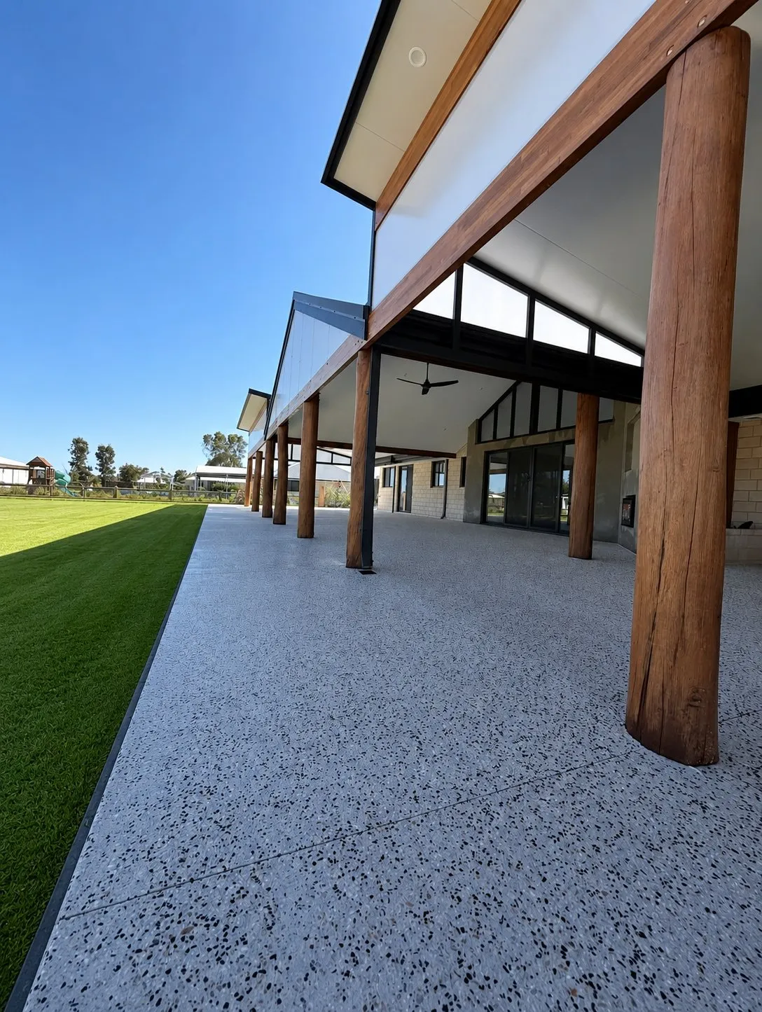Wide exposed aggregate alfresco running the length of a home under a raked ceiling with timber posts, opening onto green lawn