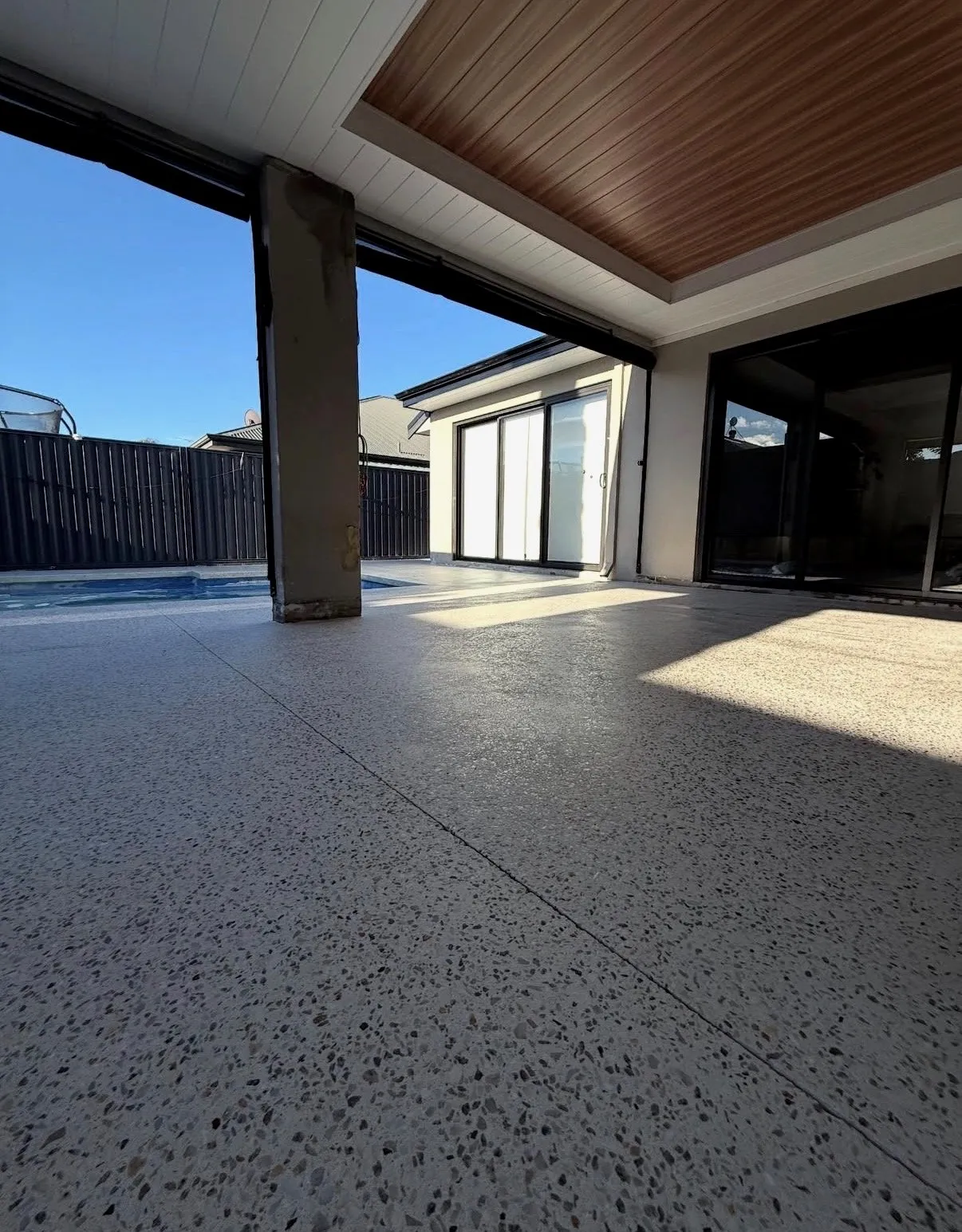 Pale exposed aggregate alfresco running from under a timber-lined patio ceiling out to a pool, with saw-cut control joints across the slab