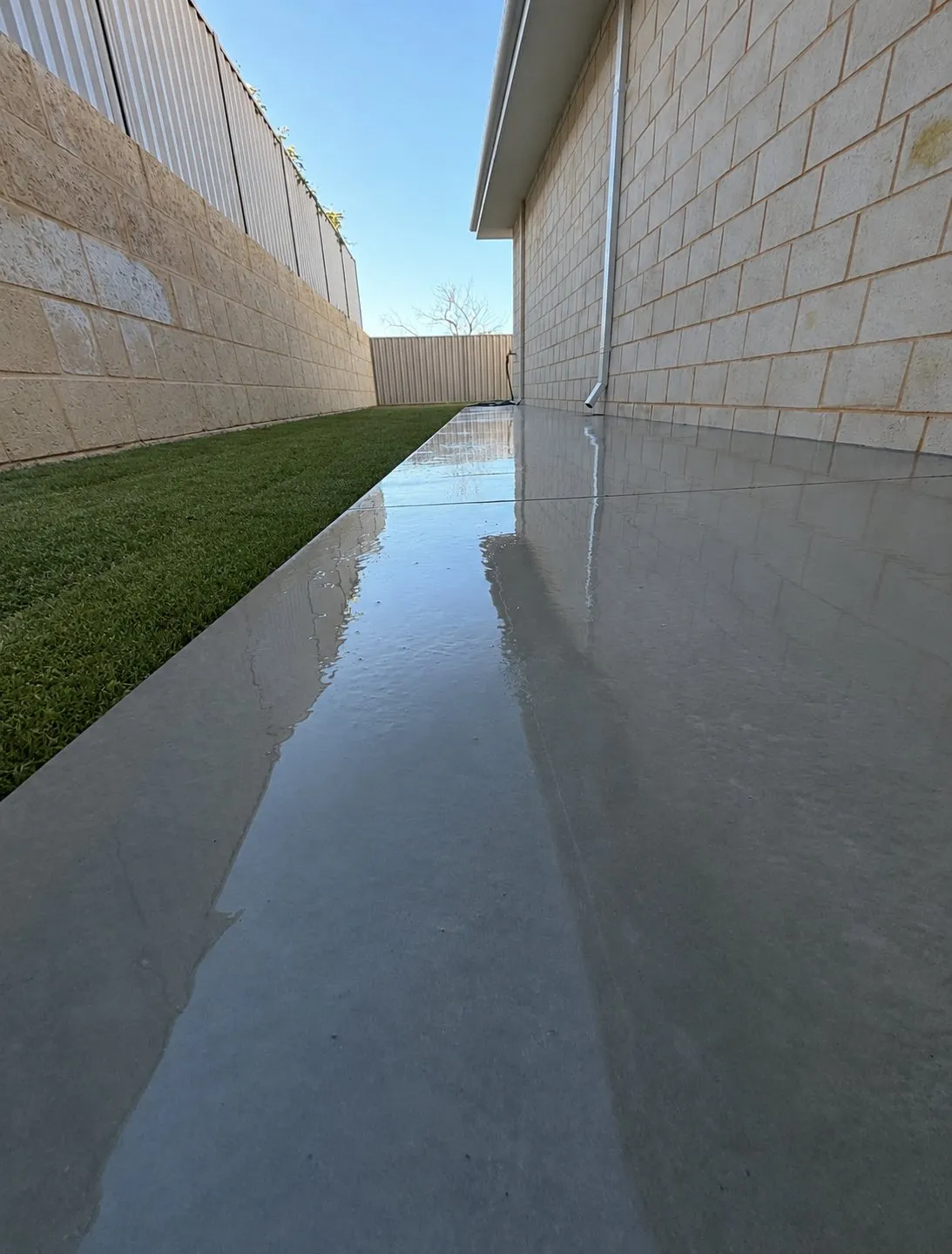 Smooth grey trowel-finished concrete path running between a limestone-block wall and a rendered house wall, reflecting the sky after rain