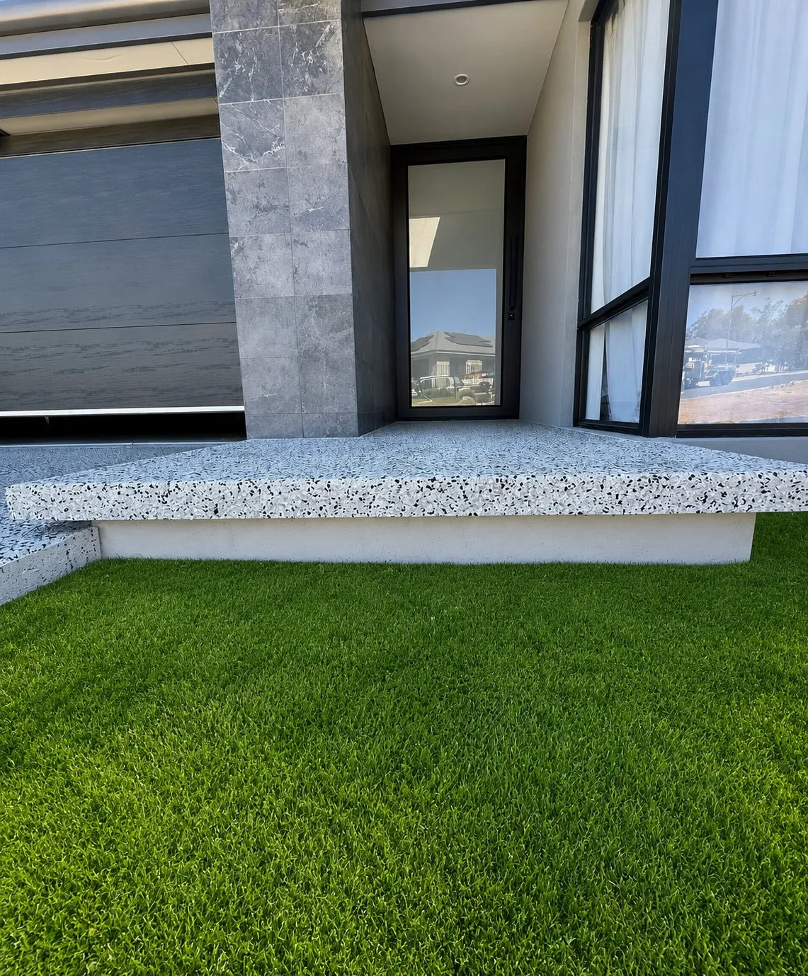 Honed exposed aggregate entry platform with a crisp cast edge, set against a marble-clad column and dark garage door with artificial turf in front