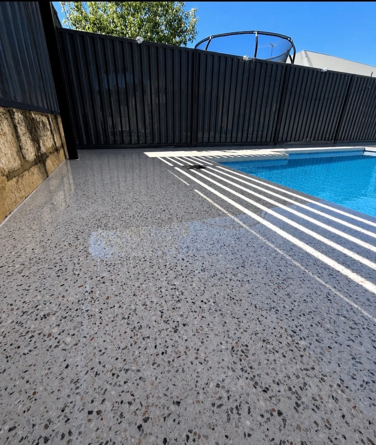 Pale speckled exposed aggregate surround beside a blue pool, striped with shadow from a dark boundary fence