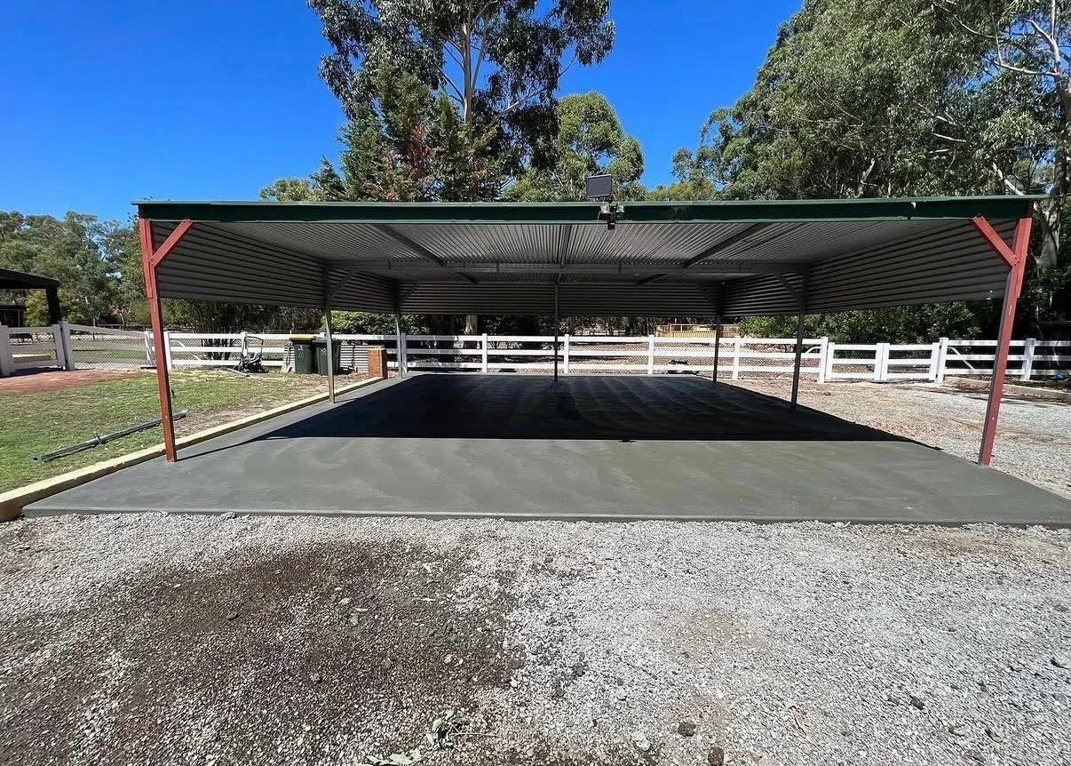 Concrete slab poured under an open-sided steel shed with a green roof, on a rural block with white post-and-rail fencing