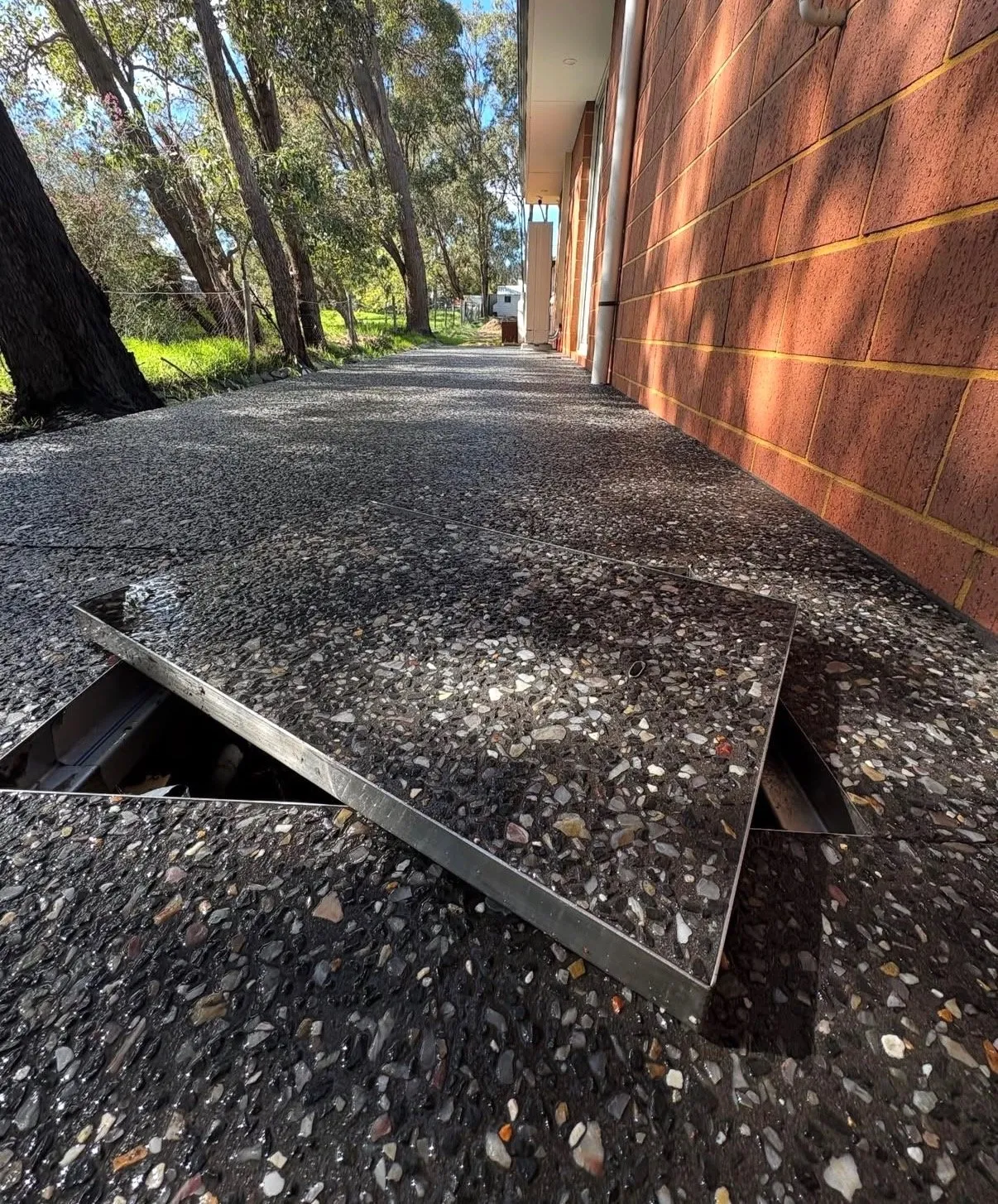 Dark charcoal exposed aggregate pathway beside a brick wall, with flush stainless steel access covers set into the surface