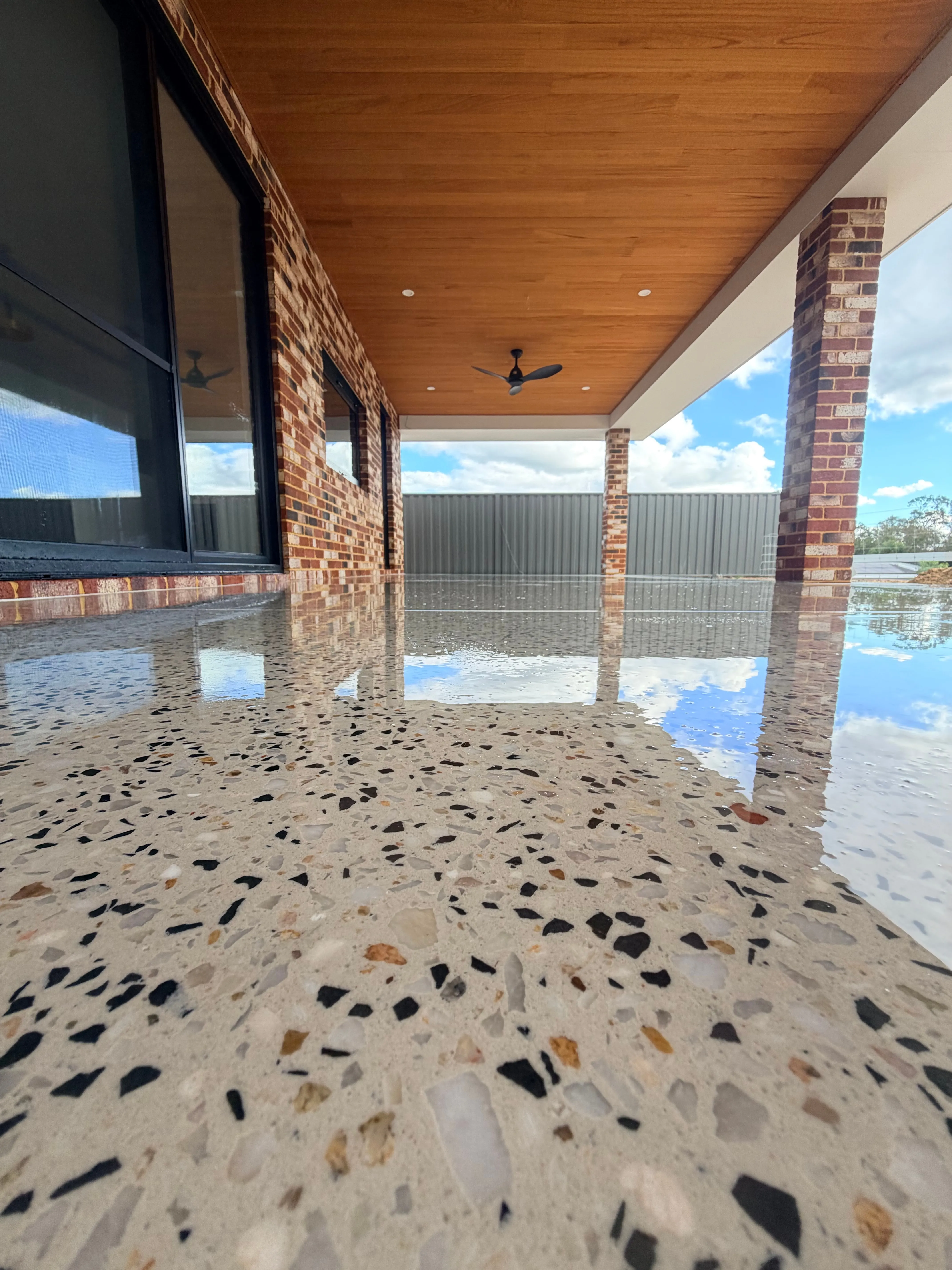 Honed concrete alfresco polished to a mirror finish, reflecting the sky in surface water, under a timber-lined ceiling between red brick piers