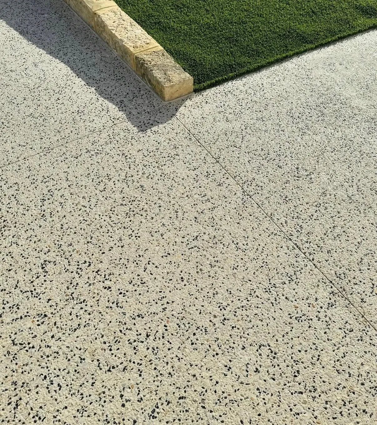 Fine white and black exposed aggregate paving with saw-cut control joints, edged with limestone blocks against artificial turf