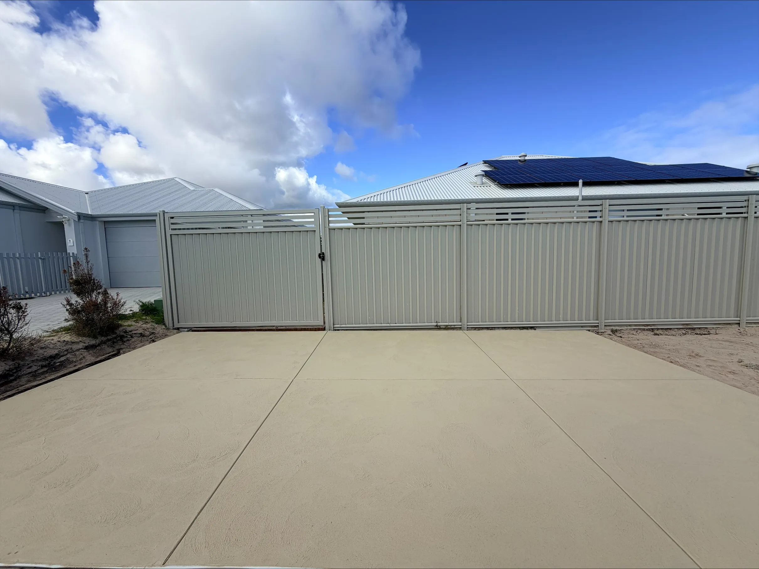 Wide sandstone-coloured concrete driveway and side access beside a new two-storey home, cream fencing and gates behind, saw-cut joints across the slab