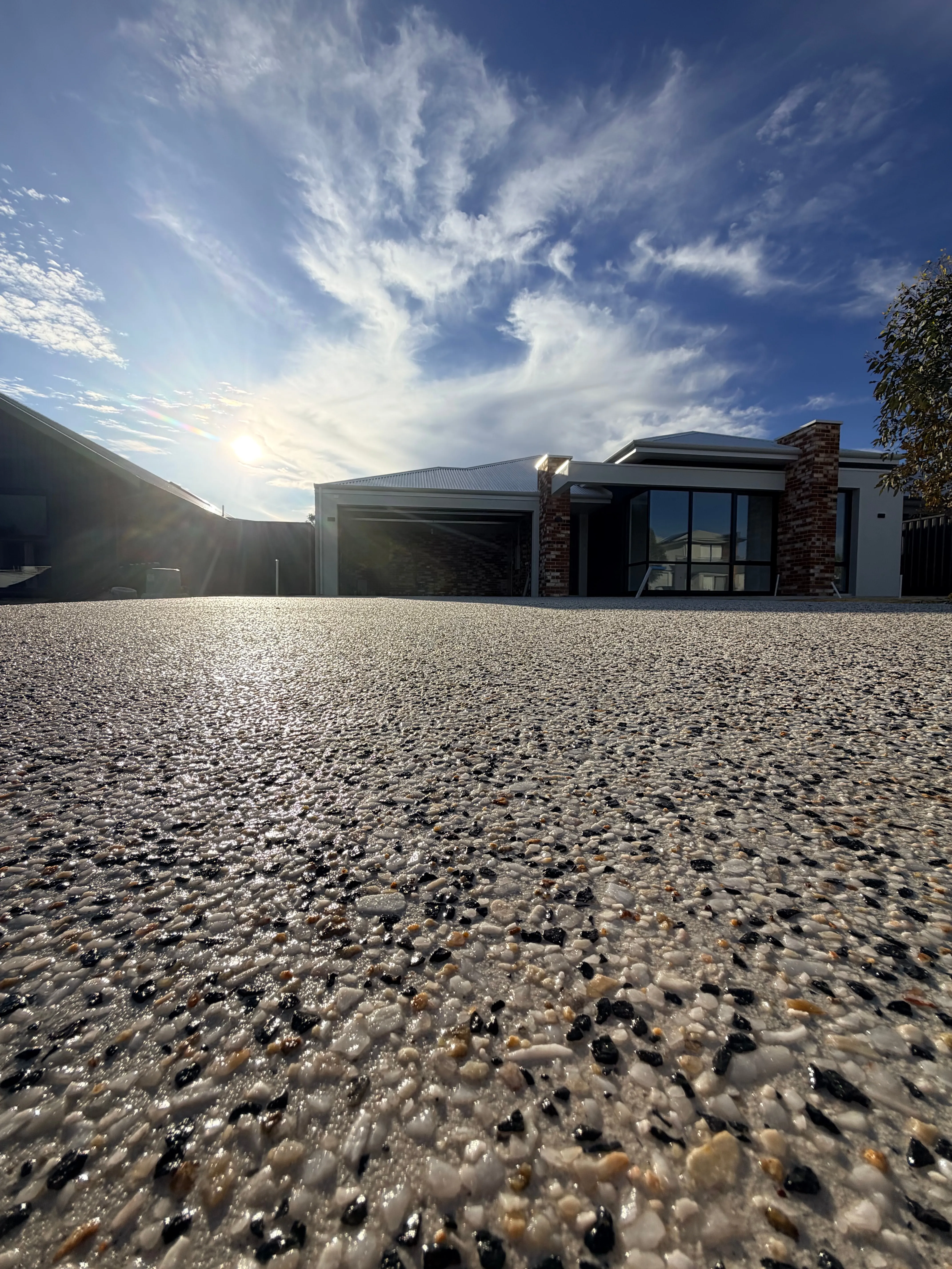 White and black exposed aggregate driveway at a modern render and brick home, lit by a low sun with a wide cirrus sky above