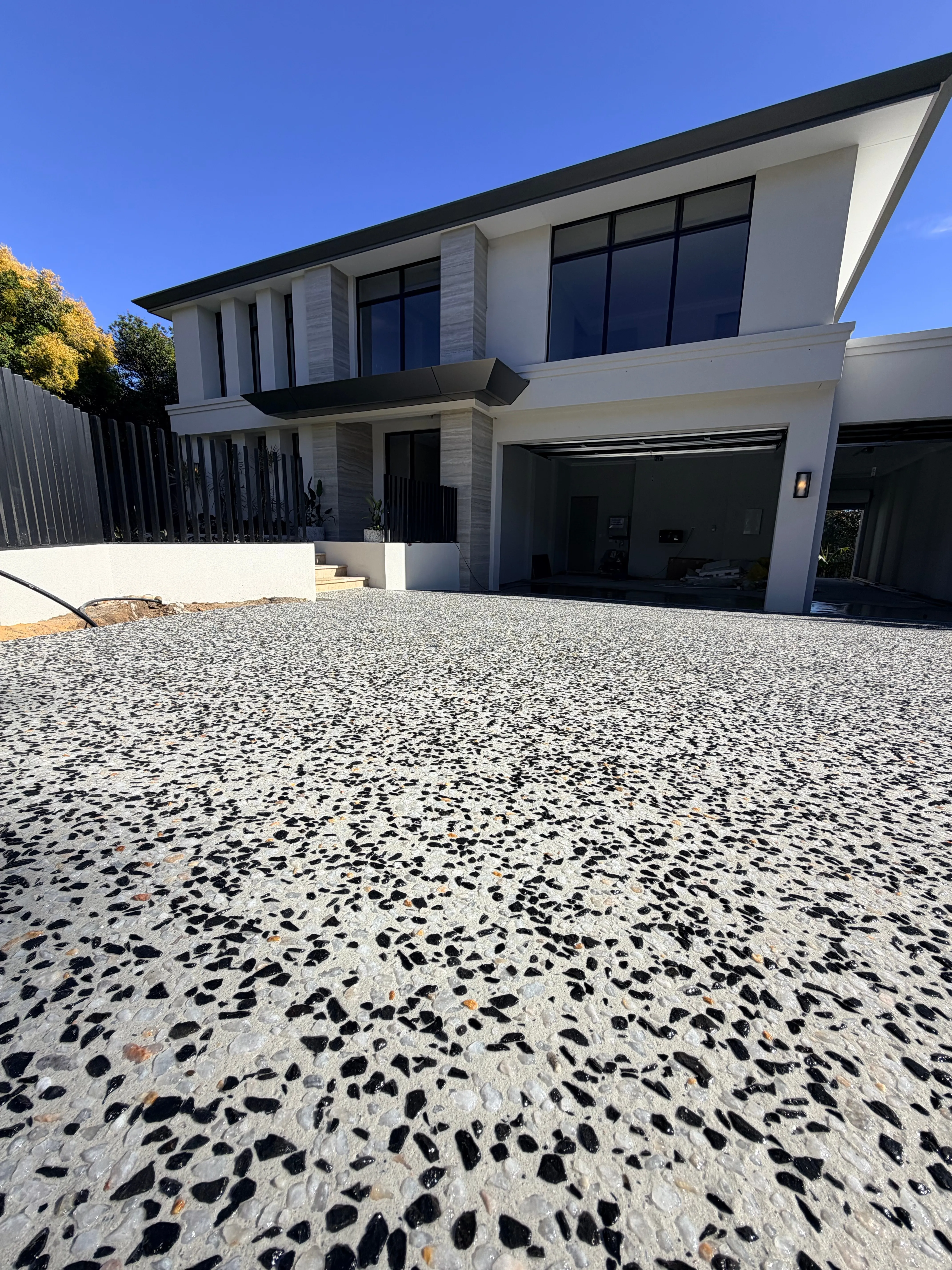 Black and white exposed aggregate driveway sweeping up to the garages of a large two-storey render and stone home