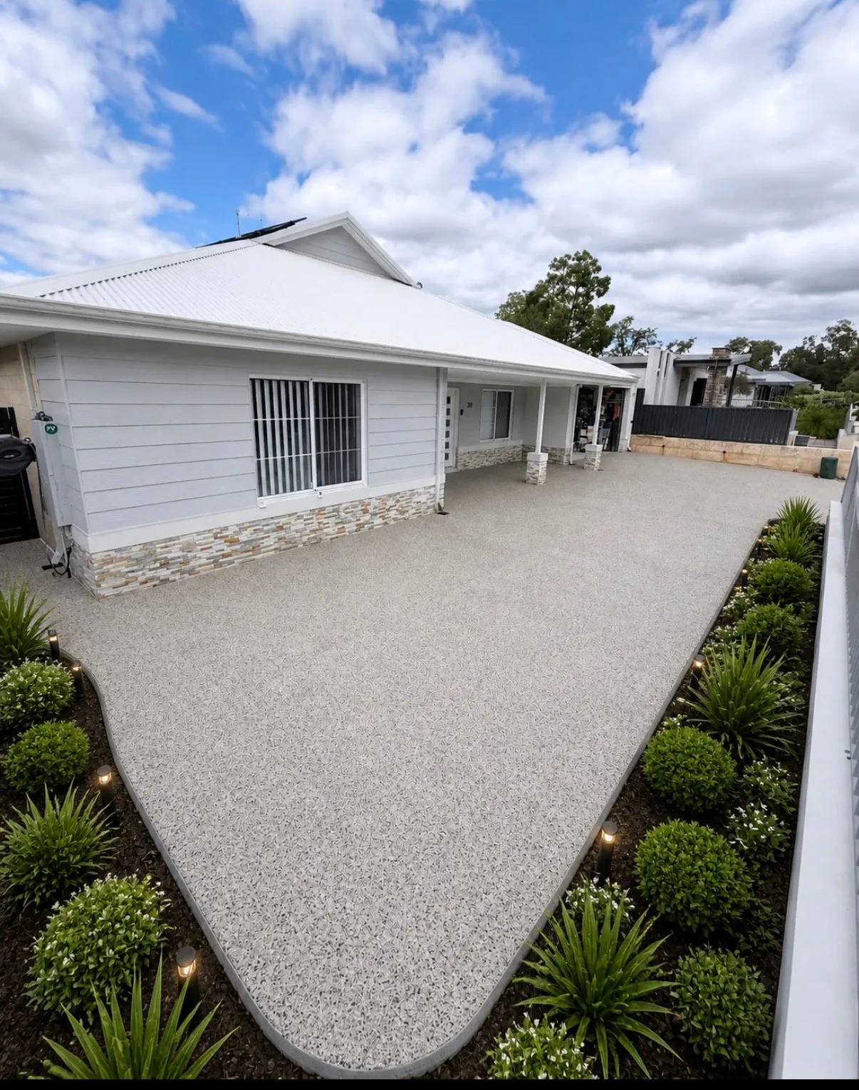 Pale cream exposed aggregate driveway curving up to a white weatherboard home with a stacked stone base, edged by garden beds and uplighting