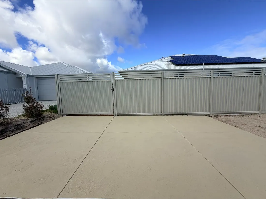 Wide sandstone-coloured concrete driveway and side access beside a new two-storey home, cream fencing behind and saw-cut joints across the slab