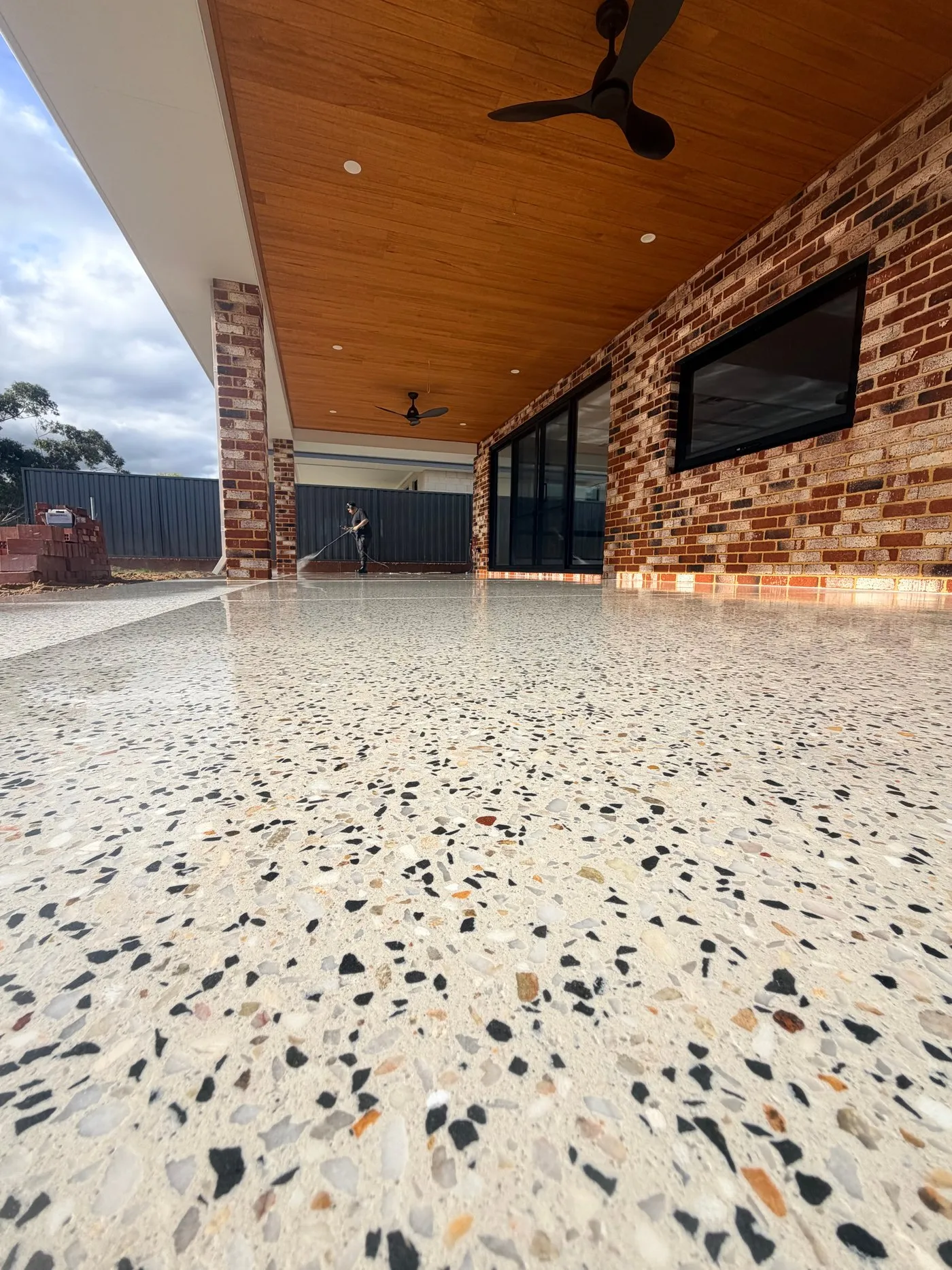 Honed concrete alfresco under a timber-lined ceiling with red brick piers, the polished floor reflecting the sky