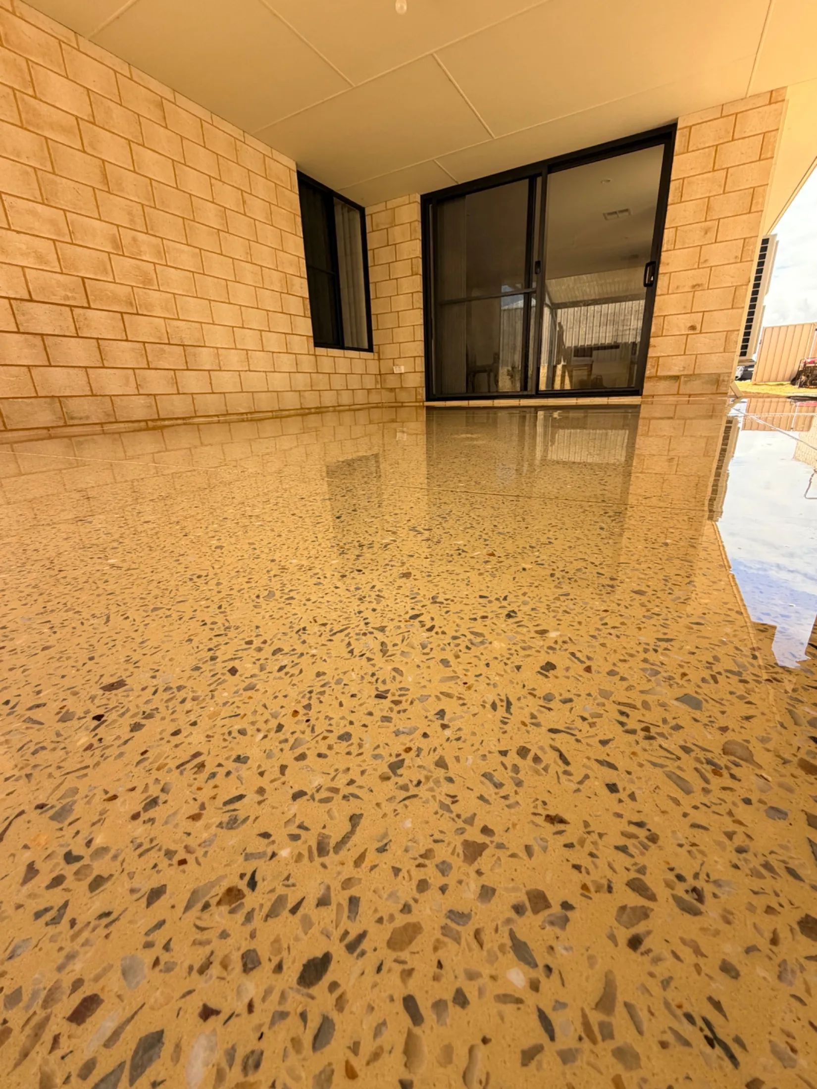 Close, low view of the Karnup honed concrete showing grey, white and amber stone in a cream base, running back to a black-framed sliding door and window in a cream brick wall