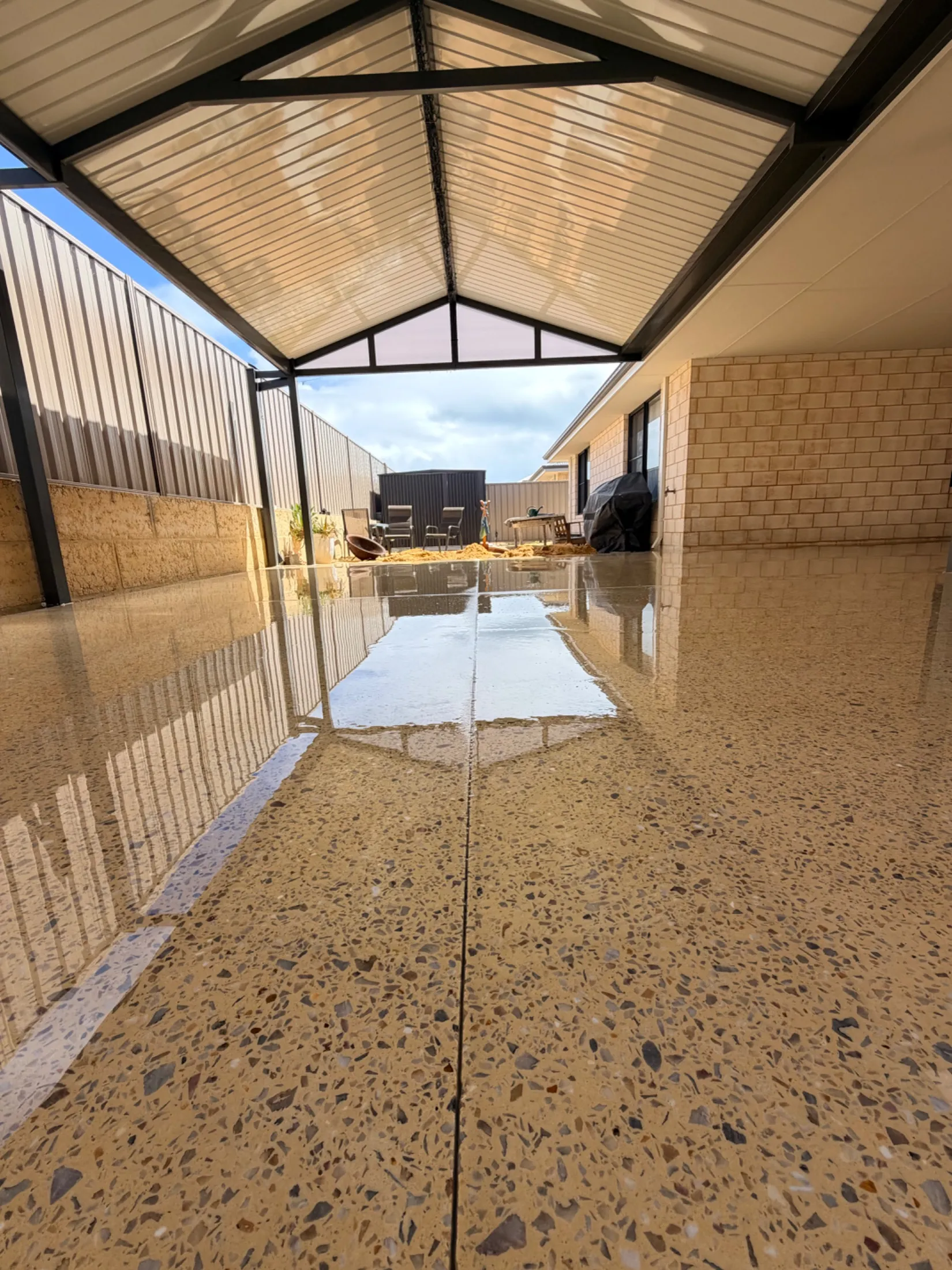 Honed concrete alfresco in Karnup shot from floor level under a gable patio roof, a saw-cut joint running down the middle and the side fence reflected in the glossy surface