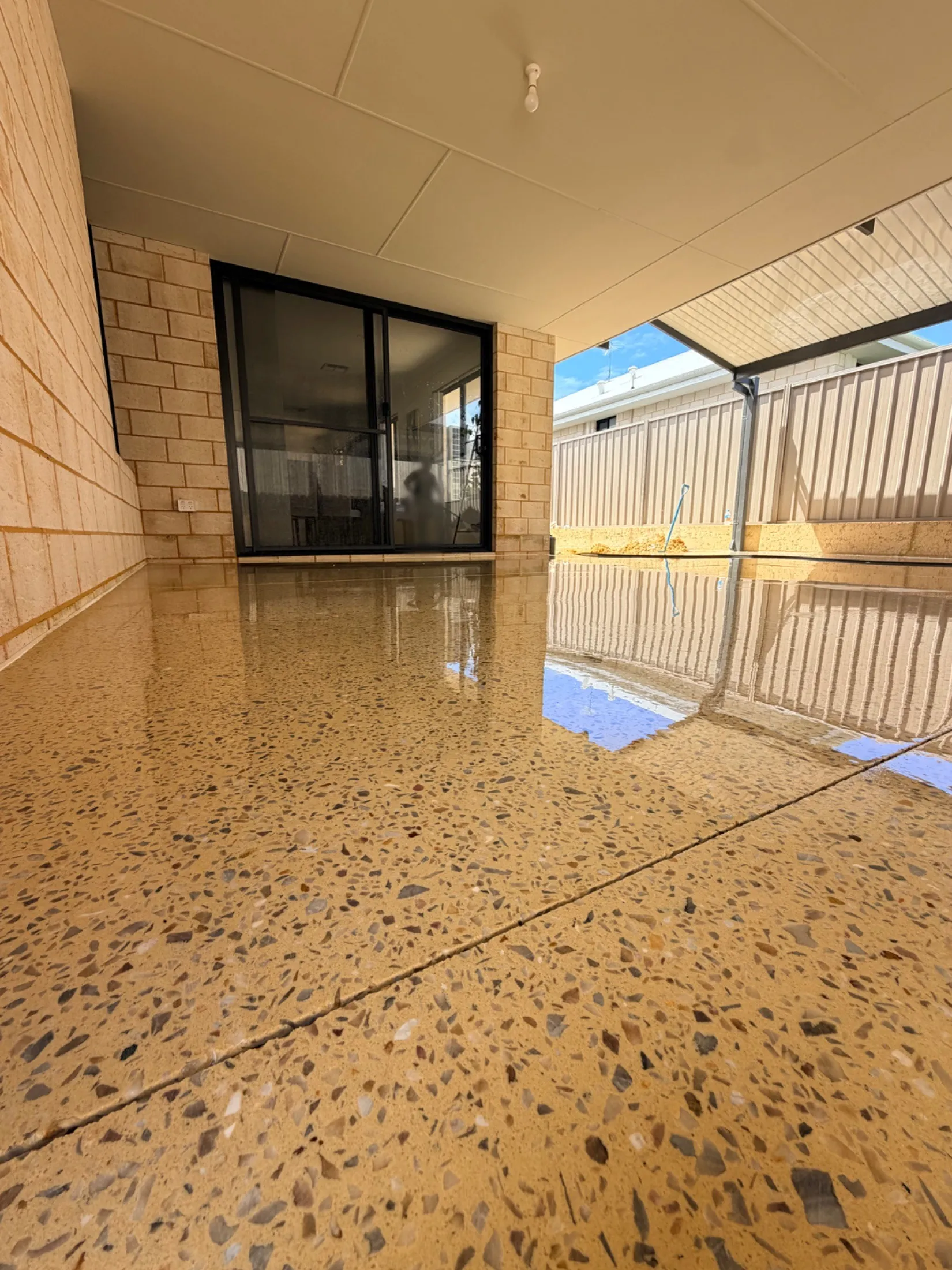 Honed concrete alfresco in Karnup meeting a sliding door under the main roof, with a saw-cut joint crossing the slab and the fence and sky reflected in the surface