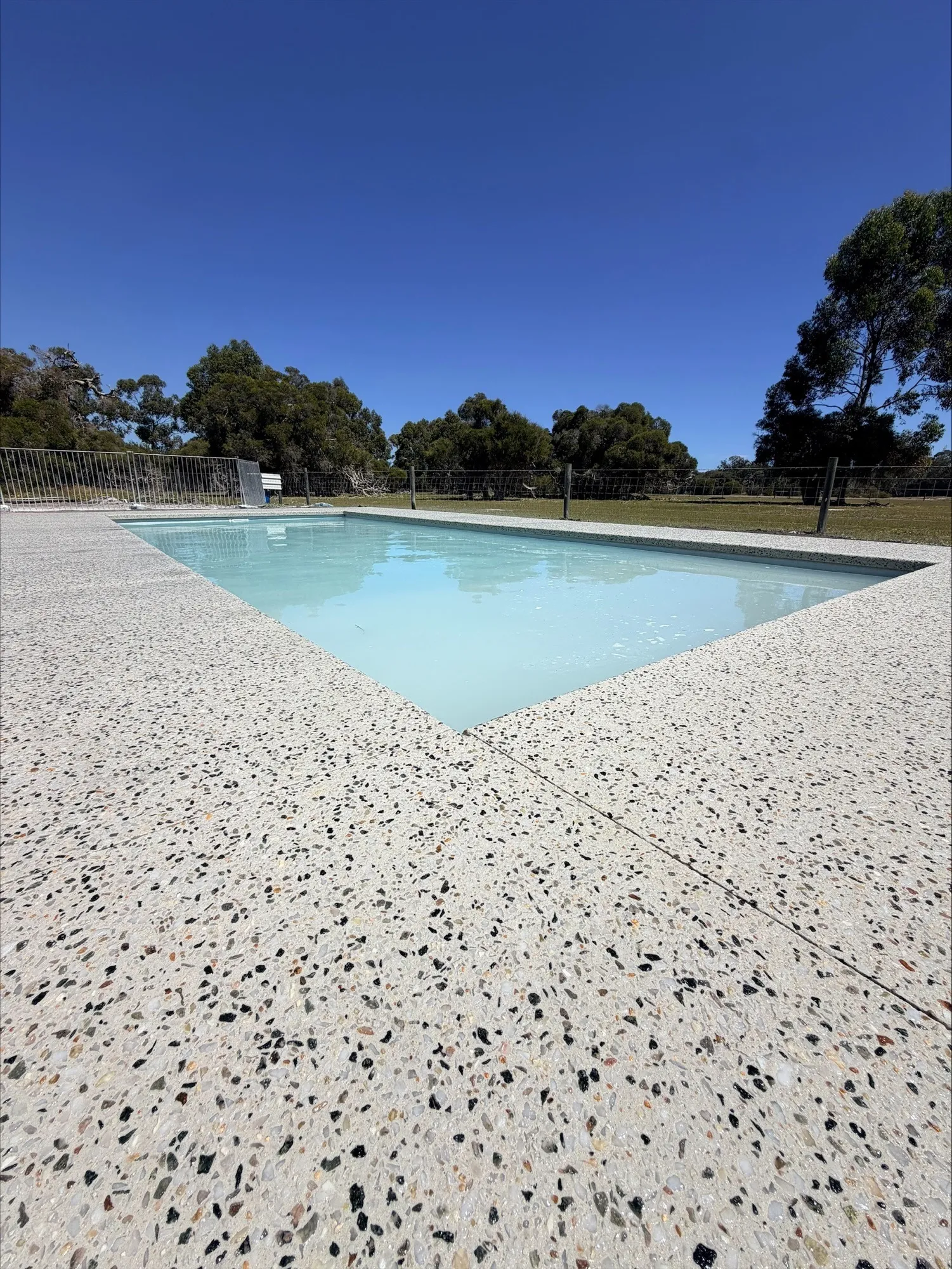 Corner of the North Dandalup pool with white exposed aggregate on both sides and a saw-cut joint running diagonally across the slab, paddock and gum trees beyond the pool fence