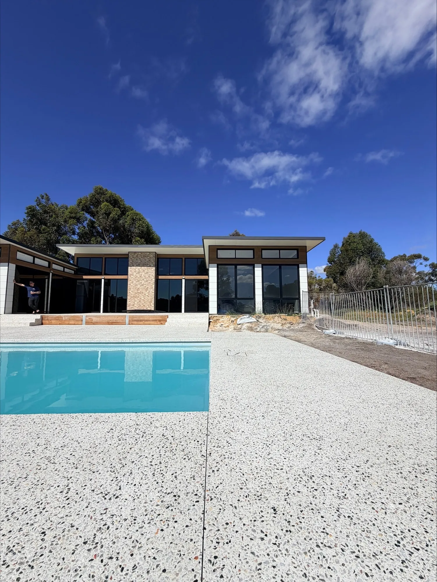 White exposed aggregate surround around an in-ground pool, running back to a modern single-storey home with a stone-clad feature wall, under a deep blue sky in North Dandalup
